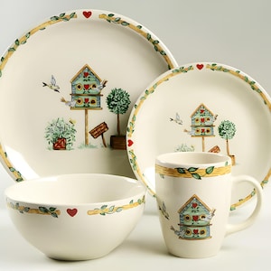 May include: A white ceramic dinnerware set with a birdhouse design. The set includes a dinner plate, salad plate, and a mug. The birdhouse is green with red hearts and is surrounded by green leaves and birds.