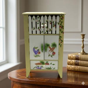 May include: A small, decorative cabinet with a light green finish. The cabinet features a white picket fence design on the top and painted vegetables on the doors and drawer. The cabinet is on a wooden table.