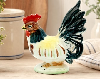 May include: A ceramic rooster figurine with a red comb, yellow chest, and green and black tail feathers. The rooster is perched on a green base. The figurine is a decorative item, likely for a kitchen or farmhouse-style decor.