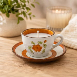 May include: A white ceramic teacup candle with a single centered wick, featuring a hand-painted orange flower with green leaves on the front. The cup sits on a matching saucer with a wide brown rim, and the interior of the cup has a brown edge.