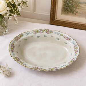 May include: Oval porcelain serving dish with a scalloped edge and floral pattern. The rim is decorated with pink, blue, and yellow flowers, and a gold trim. The dish is set on a white tablecloth.