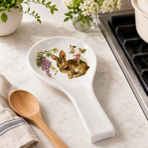 May include: A white ceramic spoon rest featuring a central illustration of a brown rabbit surrounded by purple wisteria and green foliage. The spoon rest has a wide, rounded bowl and a long, rectangular handle, designed for holding kitchen utensils.