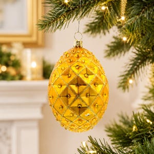 May include: A decorative, golden egg-shaped Christmas bauble with a diamond pattern and small, sparkling embellishments. It hangs from a silver hook on a Christmas tree. The background features a fireplace and soft lighting, creating a festive atmosphere.