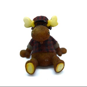 May include: A brown moose figurine wearing a red and black plaid shirt and matching cap. The moose has yellow antlers and is sitting down. The figurine is on a white background.