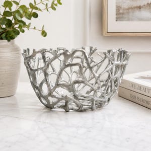 May include: A silver-colored decorative bowl featuring an intricate, open-work design that mimics the organic, branching structure of natural coral. The bowl has a metallic finish with a textured surface and a wide, rounded, open-top shape.