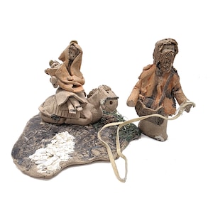 May include: Handmade ceramic nativity scene with three figures. One figure rides a donkey, another figure stands holding a strap, and a third figure is on the donkey. The figures are in shades of brown and tan, with a textured base.