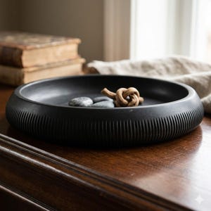 May include: A black, round decorative bowl with a textured, ribbed exterior. Inside the bowl are two gray stones and a decorative wooden knot. The bowl is sitting on a dark wooden surface.