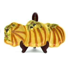 Puede incluir: Una fuente decorativa de tres secciones con forma de calabaza. La fuente está pintada de amarillo con rayas verticales marrones y detalles de hojas y bellotas verdes y marrones. La fuente se exhibe sobre un soporte marrón.