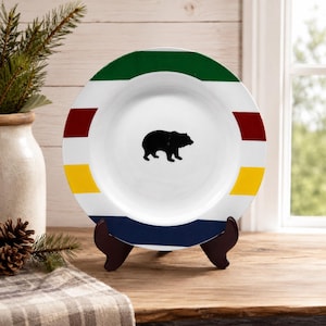 Peut inclure: Une assiette en céramique blanche avec une silhouette d'ours noir au centre. L'assiette présente un motif rayé avec des bandes vertes, rouges, jaunes et bleues. Elle est présentée sur un support en bois marron, pour la décoration intérieure.