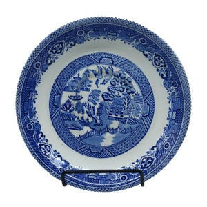 May include: A round, white and blue ceramic plate with a detailed Willow pattern design. The plate features a central scene with a bridge, trees, and buildings, surrounded by a decorative border. The plate is displayed on a black metal stand.