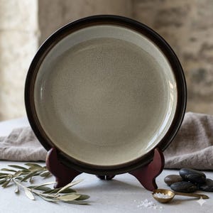 May include: A ceramic plate with a speckled, light gray center and a dark brown rim. The plate is displayed on a wooden stand. The plate is part of a tableware set.