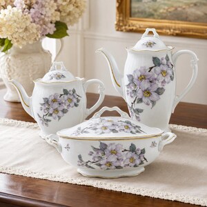 May include: A white porcelain tea set featuring a teapot, a creamer, and a covered serving dish. Each piece is decorated with a floral design in shades of purple and yellow, with gold trim. The set is arranged on a wooden table.