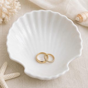 May include: A white, seashell-shaped dish holds two gold rings. One ring is a simple band, and the other is adorned with small, sparkling stones. The dish is on a neutral-toned surface, with coral and a starfish nearby.