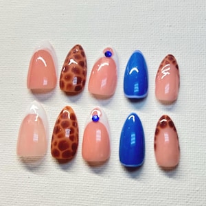 May include: A set of ten almond-shaped artificial nails. The nails feature a variety of designs, including a French tip, leopard print, solid blue, and a pink base with a small blue gem. The nails are glossy and appear to be professionally done.