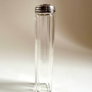 Puede incluir: Una botella de vidrio transparente con una tapa plateada. La botella tiene nervaduras verticales y un fondo redondeado.