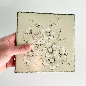 May include: A square ceramic tile with a pale sage green background featuring a black and white floral illustration. The design depicts a cluster of five-petaled flowers with detailed line work and a distressed, dark, textured border edge.