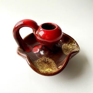 May include: A vintage ceramic ashtray with a built-in holder. The ashtray is a deep brown with yellow and brown speckles. The holder is a vibrant red with a handle. The piece has a glossy finish.