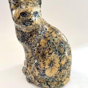May include: A ceramic cat figurine in a seated position, featuring a glossy finish with an intricate all-over floral pattern in shades of cream, muted blue, and dark grey. The cat has bright blue eyes and pointed ears, with a smooth, rounded form.