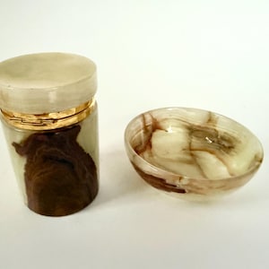 May include: A decorative jar with a lid and a small bowl. The jar is cylindrical with a gold band and a light-colored lid. The jar's body has a brown and cream-colored pattern. The bowl has a similar pattern.