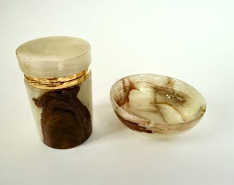 Puede incluir: Un tarro decorativo con tapa y un cuenco pequeño. El tarro es cilíndrico con una banda dorada y una tapa de color claro. El cuerpo del tarro tiene un patrón marrón y crema. El cuenco tiene un patrón similar.