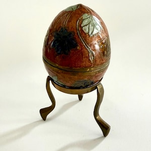 May include: An egg-shaped container with a reddish-brown enamelled surface featuring dark blue and light green floral motifs. The object is supported by a three-legged brass stand with curved feet, showcasing a metallic finish and a horizontal seam.
