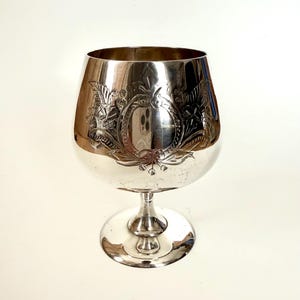 Könnte beinhalten: Ein silberfarbenes Brandyglas mit einem verzierten floralen und ovalen Design. Das Glas hat eine breite Schale und einen kurzen Stiel mit rundem Fuß. Die detaillierte Gravur verleiht diesem Vintage-Glas eine elegante Note.