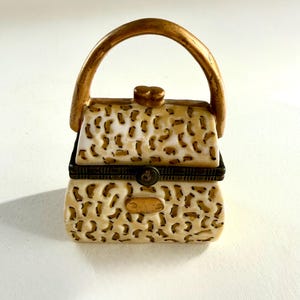 May include: A small, decorative trinket box shaped like a handbag with a cream-coloured body featuring a brown leopard print pattern. It includes a gold-toned arched handle, a central gold-toned clasp, and a dark metal hinge running across the middle.