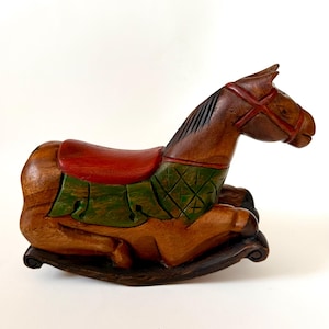 May include: A wooden rocking horse, hand-carved and stained brown. It has a red saddle and a green blanket with a diamond pattern. The bridle is red. The toy is designed for decorative purposes.