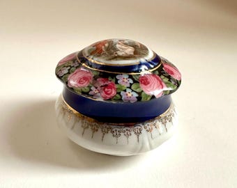 May include: A small, round porcelain trinket box with a blue and gold lid. The lid is decorated with a floral pattern and a painted scene of two figures. The box is perfect for storing small treasures.