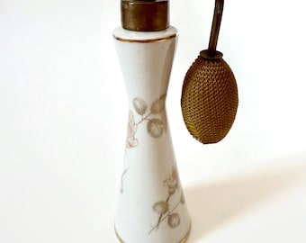 May include: A vintage perfume bottle. The white porcelain body has a floral design in soft pink and gold accents. It features a brass atomizer and a gold-trimmed base, creating an elegant, antique aesthetic.