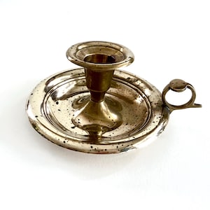 May include: A polished brass chamberstick candle holder featuring a wide, circular drip tray with a speckled patina finish. The central socket is designed to hold a single taper candle, and a small, circular finger loop handle is attached to the side.