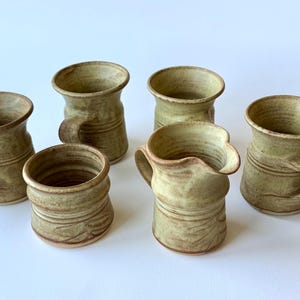 May include: Six handmade ceramic mugs and a small pitcher in a light green and brown color palette. The mugs have a rounded shape with a handle and a textured surface. The pitcher has a spout.