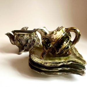 May include: A green and brown seashell-shaped teapot, creamer, and four matching plates. The teapot and creamer have a metal handle.