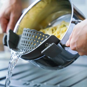 Könnte beinhalten: Ein Topf aus Edelstahl mit einem grauen Silikoneinsatz. Der Topf wird über einem Waschbecken gehalten und Wasser läuft aus dem Topf.