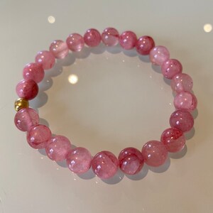 Puede incluir: Un brazalete de piedras preciosas rosas con cuentas redondas translúcidas con inclusiones rojas. Un pequeño elemento dorado acentúa el diseño. El brazalete es una joya delicada, ideal para el uso diario.