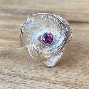 May include: A silver-colored ring with a textured, swirling design. The ring features a round, purple gemstone set in the center. The ring is displayed on a wooden surface.