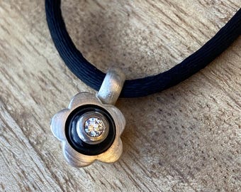 May include: A silver flower pendant necklace with a black cord. The pendant has a flower shape with a black center and a sparkling gemstone. The necklace is on a wooden surface.