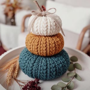 Op de afbeelding: Een stapel van drie gebreide pompoenen in crème, mosterd en blauwgroen. De pompoenen staan op een wit dienblad, met een takje en lint bovenop. Een herfstthema decoratie.
