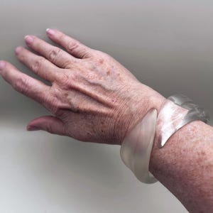 May include: Two translucent, chunky bracelets. One is a light, creamy color, and the other is a clear, frosted gray. The bracelets are worn on a wrist, showcasing their bold, geometric shapes and smooth, polished surfaces. The bracelets are a statement accessory.