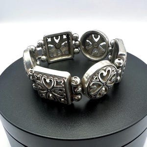 Puede incluir: Pulsera plateada con una serie de eslabones decorativos. La pulsera presenta eslabones cuadrados, ovalados y redondos, cada uno con un dise&ntilde;o floral o en forma de coraz&oacute;n. Los eslabones est&aacute;n conectados por secciones m&aacute;s peque&ntilde;as con cuentas. La pulsera se muestra sobre una superficie negra.