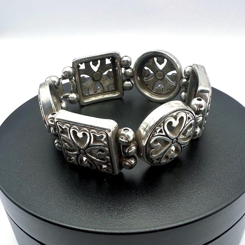 Puede incluir: Pulsera plateada con una serie de eslabones decorativos. La pulsera presenta eslabones cuadrados, ovalados y redondos, cada uno con un dise&ntilde;o floral o en forma de coraz&oacute;n. Los eslabones est&aacute;n conectados por secciones m&aacute;s peque&ntilde;as con cuentas. La pulsera se muestra sobre una superficie negra.