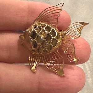 May include: A gold-colored fish-shaped brooch with intricate filigree detailing. The brooch features a detailed design of a fish with fins and a tail. The fish's body has a textured pattern, and the brooch is held in a hand.