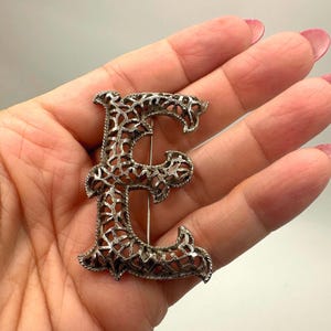 May include: Silver-toned metal letter "E" brooch. The brooch features an openwork design with intricate, swirling patterns. The pin is a decorative accessory, suitable for adding a personalized touch to clothing or accessories.