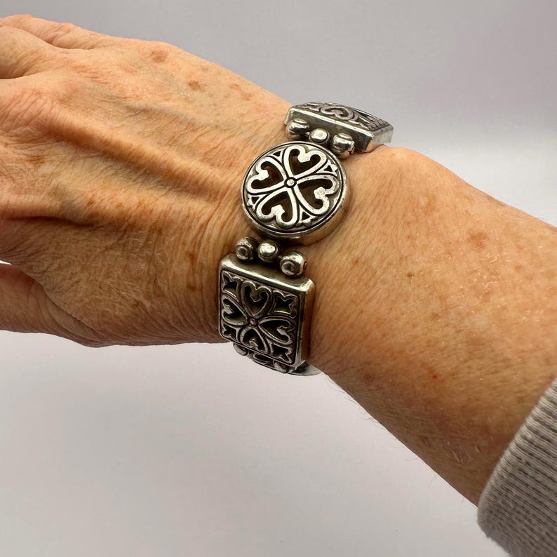 Puede incluir: Pulsera plateada con eslabones decorativos. La pulsera presenta eslabones cuadrados, redondos y rectangulares, cada uno adornado con patrones en forma de coraz&oacute;n. La pulsera est&aacute; dise&ntilde;ada para ser usada en la mu&ntilde;eca. El dise&ntilde;o es intrincado.