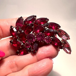 May include: A vintage brooch featuring a cluster of ruby-red and pink marquise-cut and round-cut rhinestones. The brooch has a silver-tone metal setting and is held in a hand.