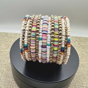 May include: A stack of colorful beaded bracelets. The bracelets feature a mix of white, gold, and multi-colored beads. The beads are arranged in a stacked design, creating a bold and eye-catching accessory. The bracelets are displayed on a black surface.