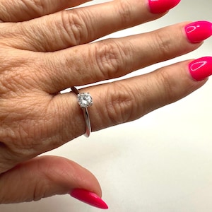 May include: A silver ring with a round, clear gemstone. The ring is on a finger with bright pink nail polish. The ring's band is thin and polished, and the gemstone is set in a classic four-prong setting. The ring is a simple and elegant piece of jewelry.