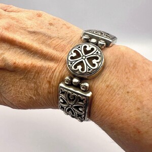 Puede incluir: Pulsera plateada con un dise&ntilde;o floral decorativo. La pulsera presenta una pieza central redonda y piezas finales cuadradas, todas con un patr&oacute;n de coraz&oacute;n calado. La pulsera es de metal y tiene un estilo vintage o antiguo.