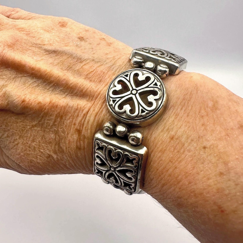 Puede incluir: Pulsera plateada con un dise&ntilde;o floral decorativo. La pulsera presenta una pieza central redonda y piezas finales cuadradas, todas con un patr&oacute;n de coraz&oacute;n calado. La pulsera es de metal y tiene un estilo vintage o antiguo.