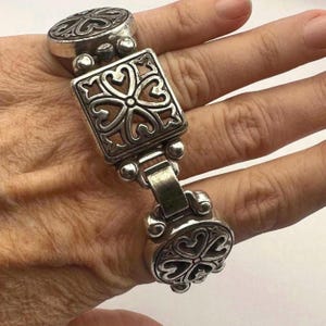 Puede incluir: Una pulsera plateada con un dise&ntilde;o ornamentado. La pulsera presenta una pieza central cuadrada con un patr&oacute;n en forma de coraz&oacute;n, conectada por un cierre articulado a extremos ovalados. El metal tiene un aspecto ligeramente envejecido.